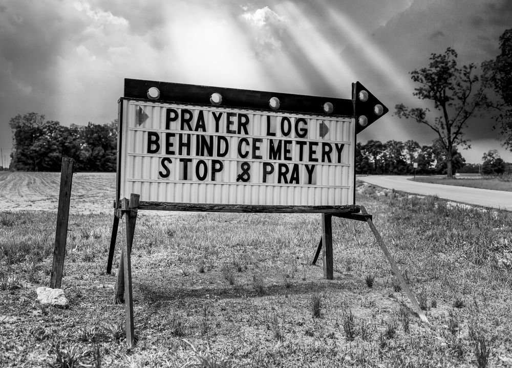 Black and white photo of a roadside sign reading Prayer Log Behind Cemetery Stop and Pray under dramatic sunbeams.
