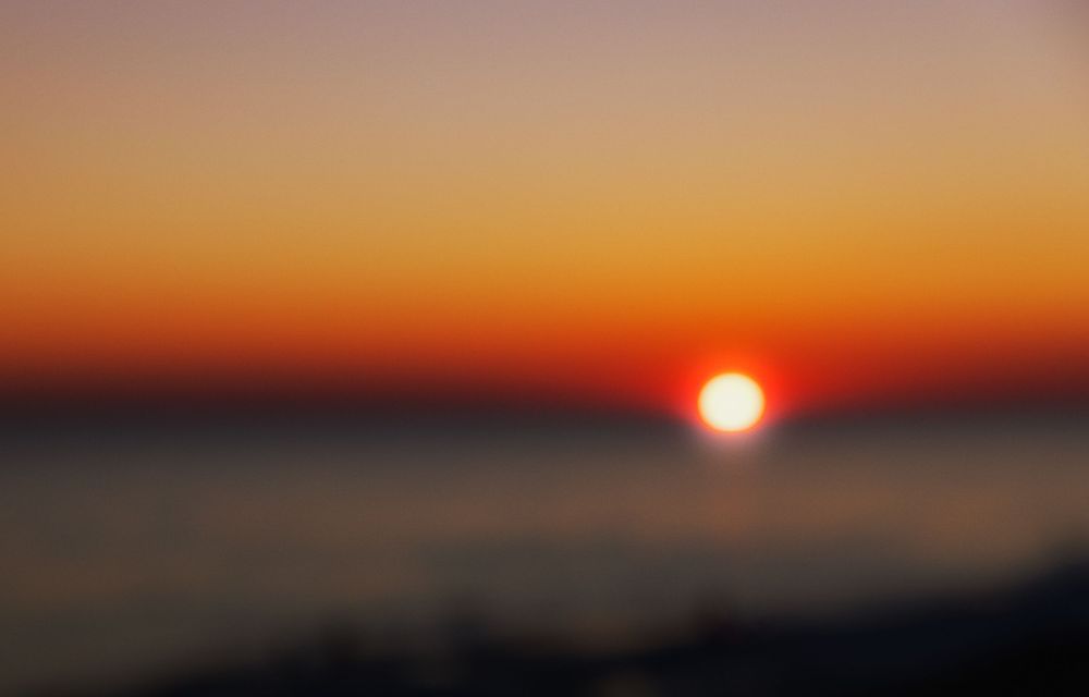 Blurry sunset over calm ocean waters with vibrant orange and red sky gradient near the horizon.