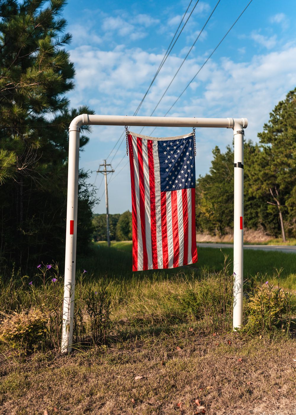 American flag hanging from white metal poles in a rural setting with power lines and trees.