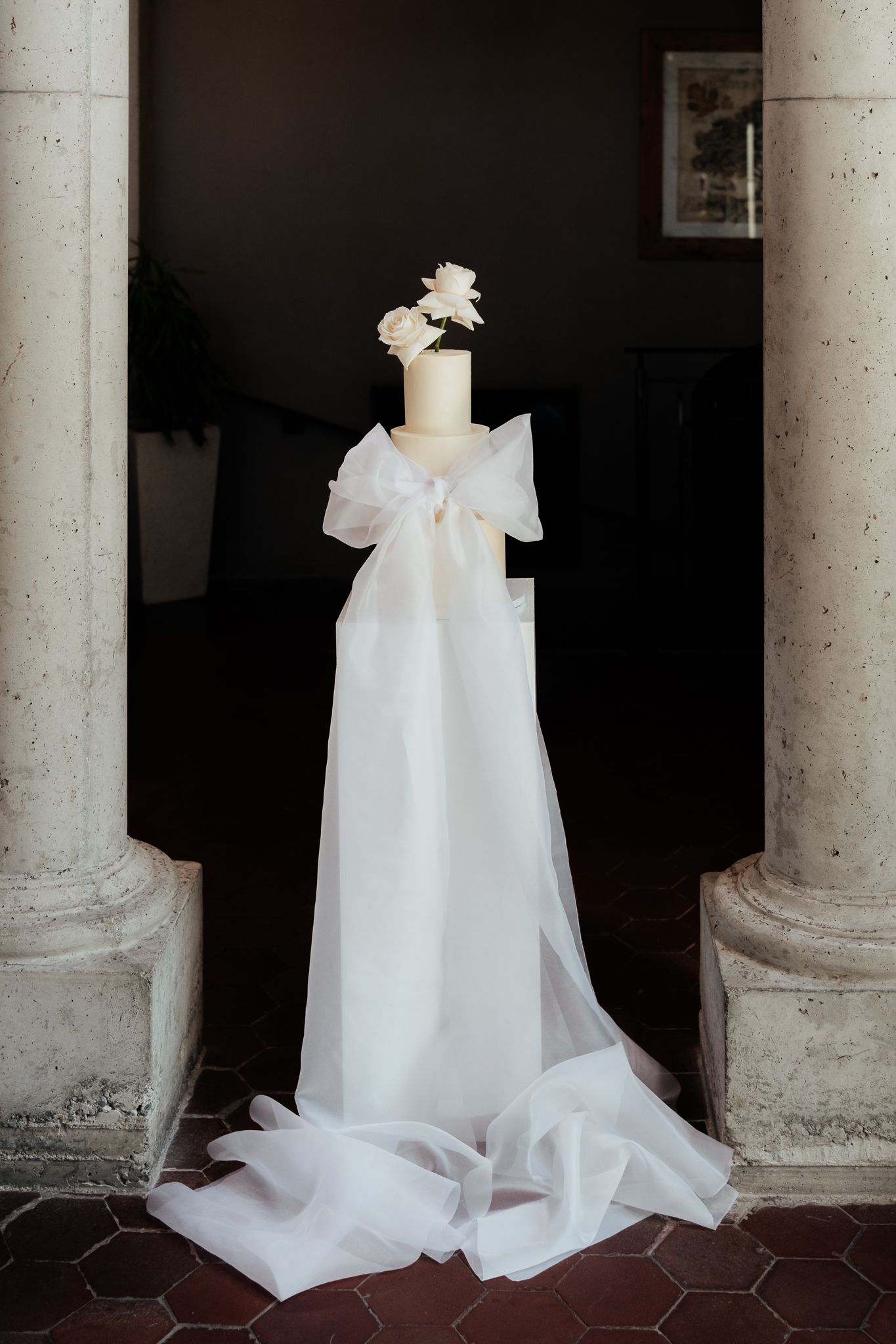 A modern white wedding cake with two white roses displayed against stone columns at the Milk Station by wedding photographer August and Vine