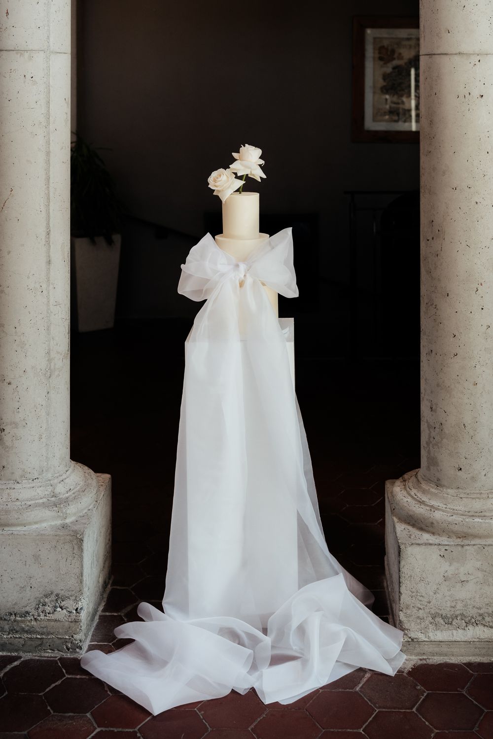 A modern white wedding cake with two white roses displayed against stone columns at the Milk Station by wedding photographer August and Vine