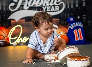 Baby wearing baseball jersey sits on table near decorative neon signs and rookie of the year backdrop.