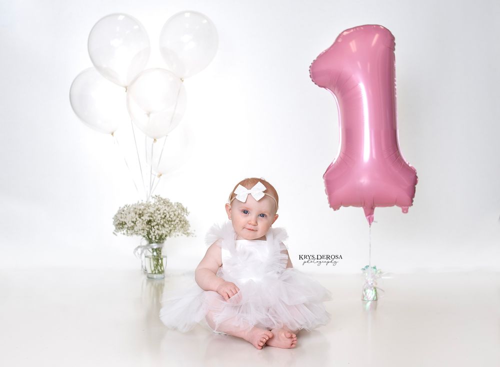A baby's first birthday photo collage with pink balloons, white flowers, and cake smash moments on light background.