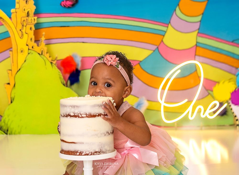 Pink frosted birthday cake with rainbow sprinkles and colorful decorations against a bright turquoise background.