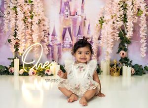 A baby in a white lace dress sits on the floor with fairy lights and castle backdrop for a magical Disney-themed photo.