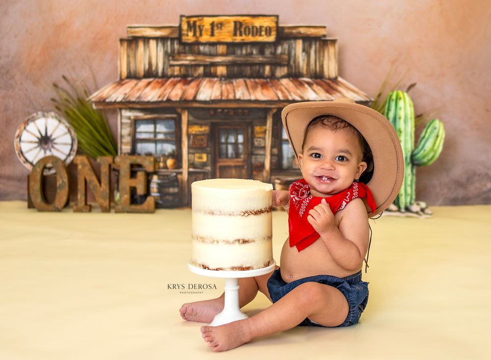 Baby in cowboy hat sits before a cake at a western-themed first birthday photo shoot with ONE sign.