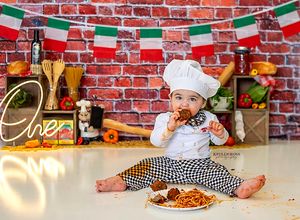 A small child in chef attire sits on the floor with pasta and Italian decor against a brick wall with hanging flags.