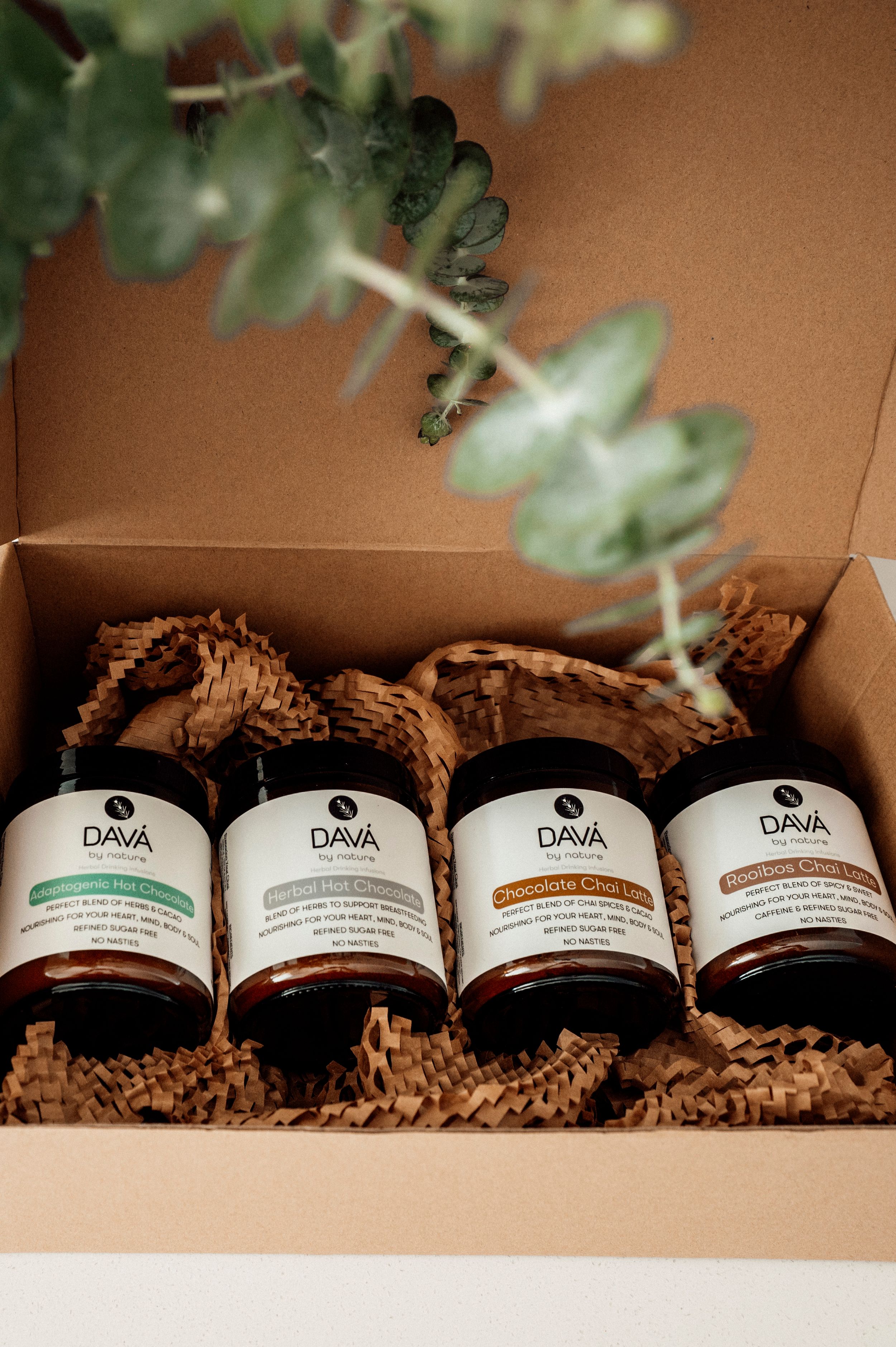 Four labeled glass jars of skincare products arranged in a brown box with decorative packing material and eucalyptus sprigs.