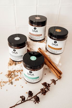 Four black-lidded glass jars of Dava brand products with cinnamon sticks and dried flowers on white tile surface.