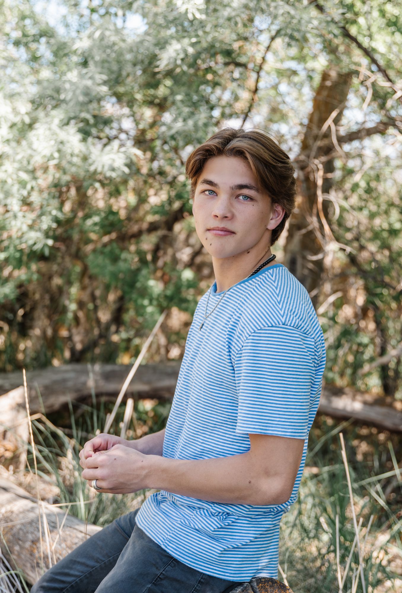 Person in striped blue shirt sitting outdoors among trees and branches.