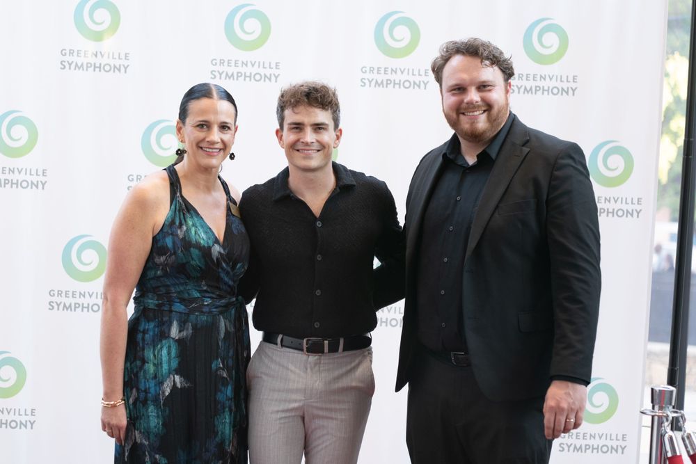 Three people pose at an event with Greenville Symphony branded backdrop.