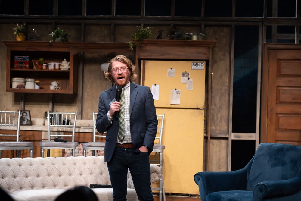 Performer speaking into microphone on stage with vintage furniture set design.