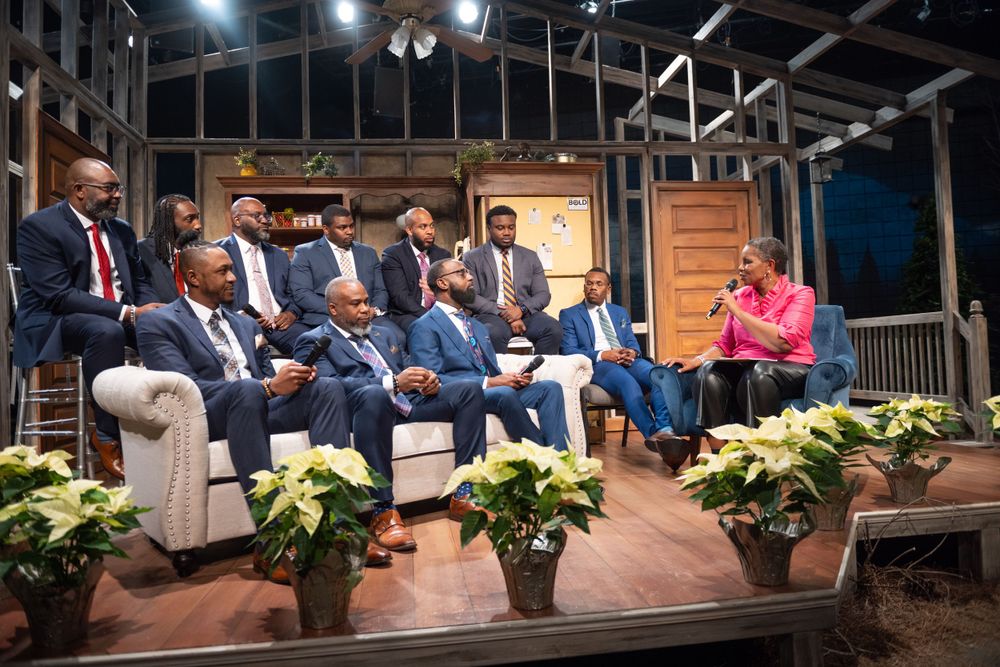 Group seated on a theatrical stage set decorated with potted plants and wooden paneling.