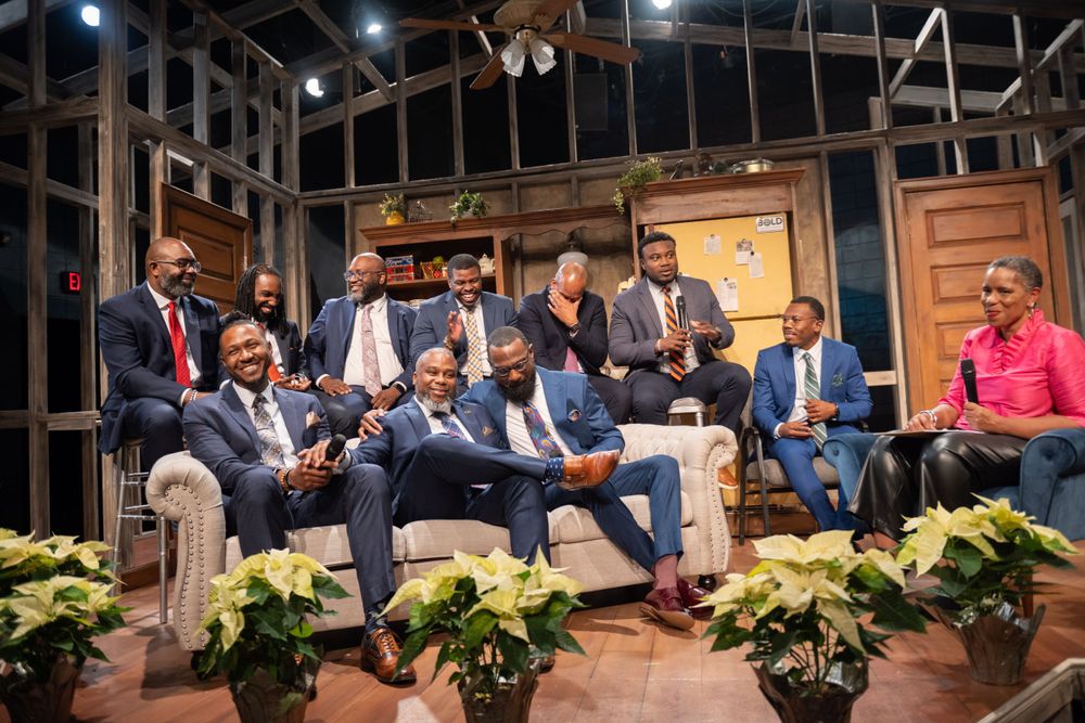 People in formal suits gathered on a rustic stage set with decorative plants.