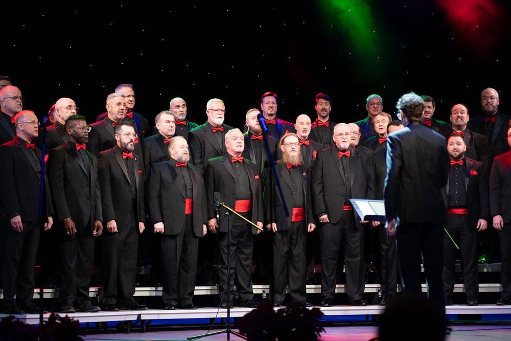 Choir ensemble in formal black attire with red accents performing on illuminated stage.