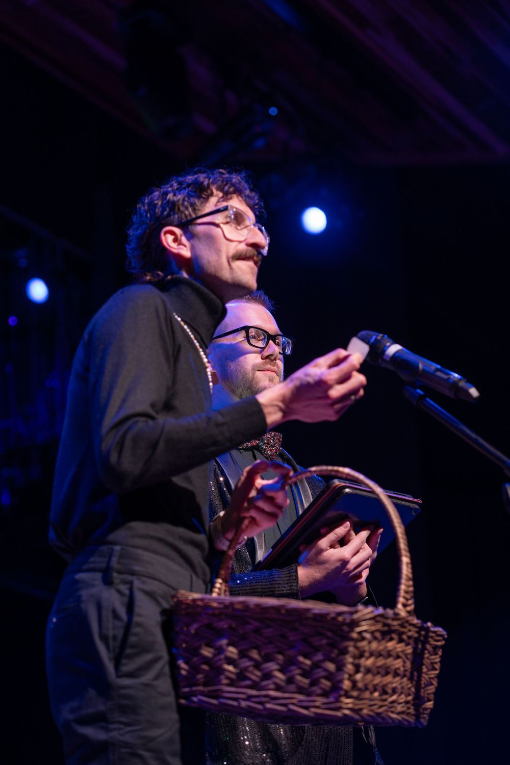 Performer in black attire holds a woven basket while speaking into microphone on dimly lit stage.