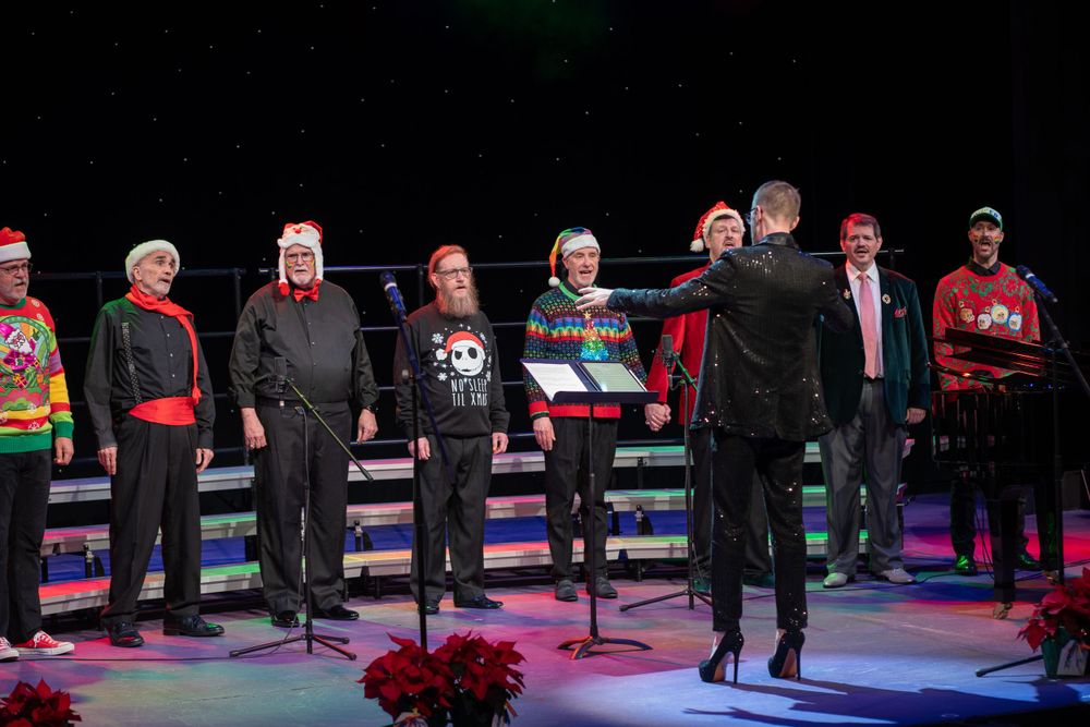 Choir performers in dark attire and holiday accessories standing on stage with red poinsettias.