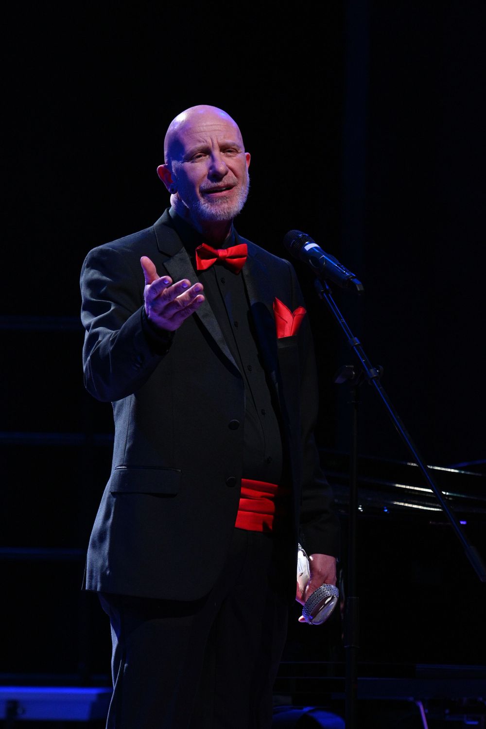 Performer in formal black suit with red accents gesturing while singing on stage.