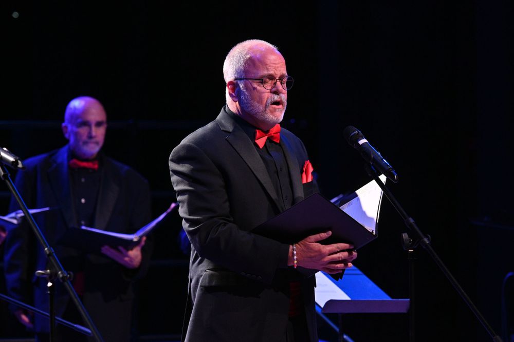 Solo performer in black suit with red bow tie singing at microphone on dark stage.
