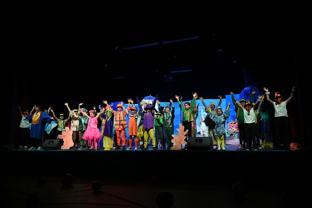 Large group in colorful costumes performing on a dark stage with blue lighting.