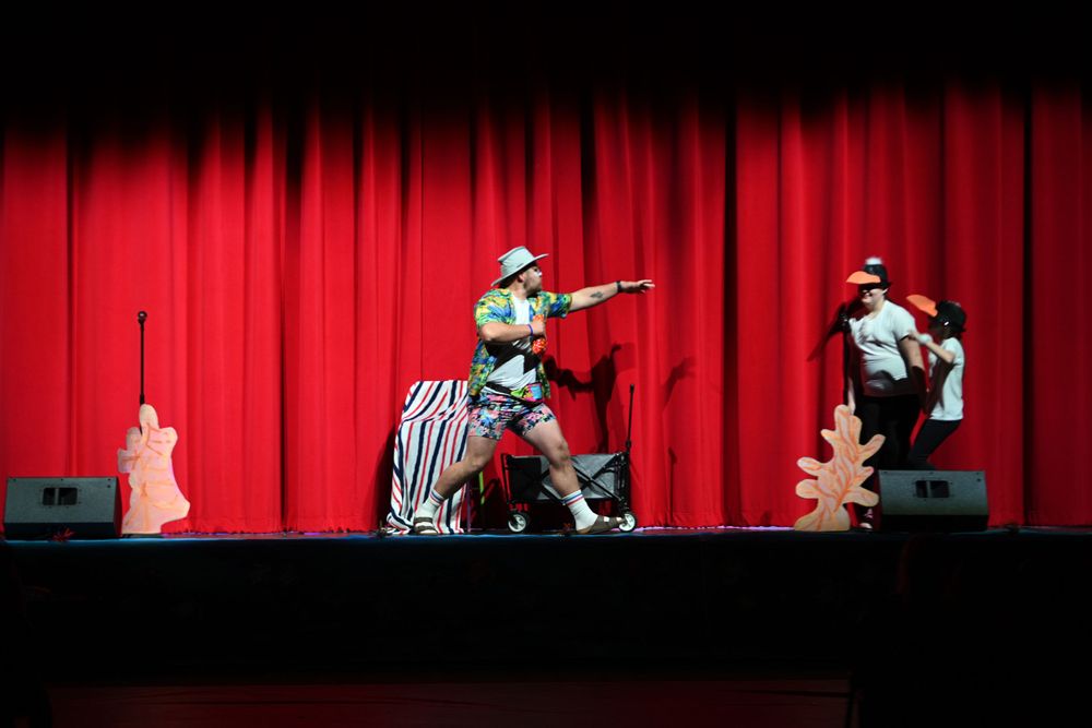 Theatrical performance on stage with performers against a dramatic red curtain backdrop.