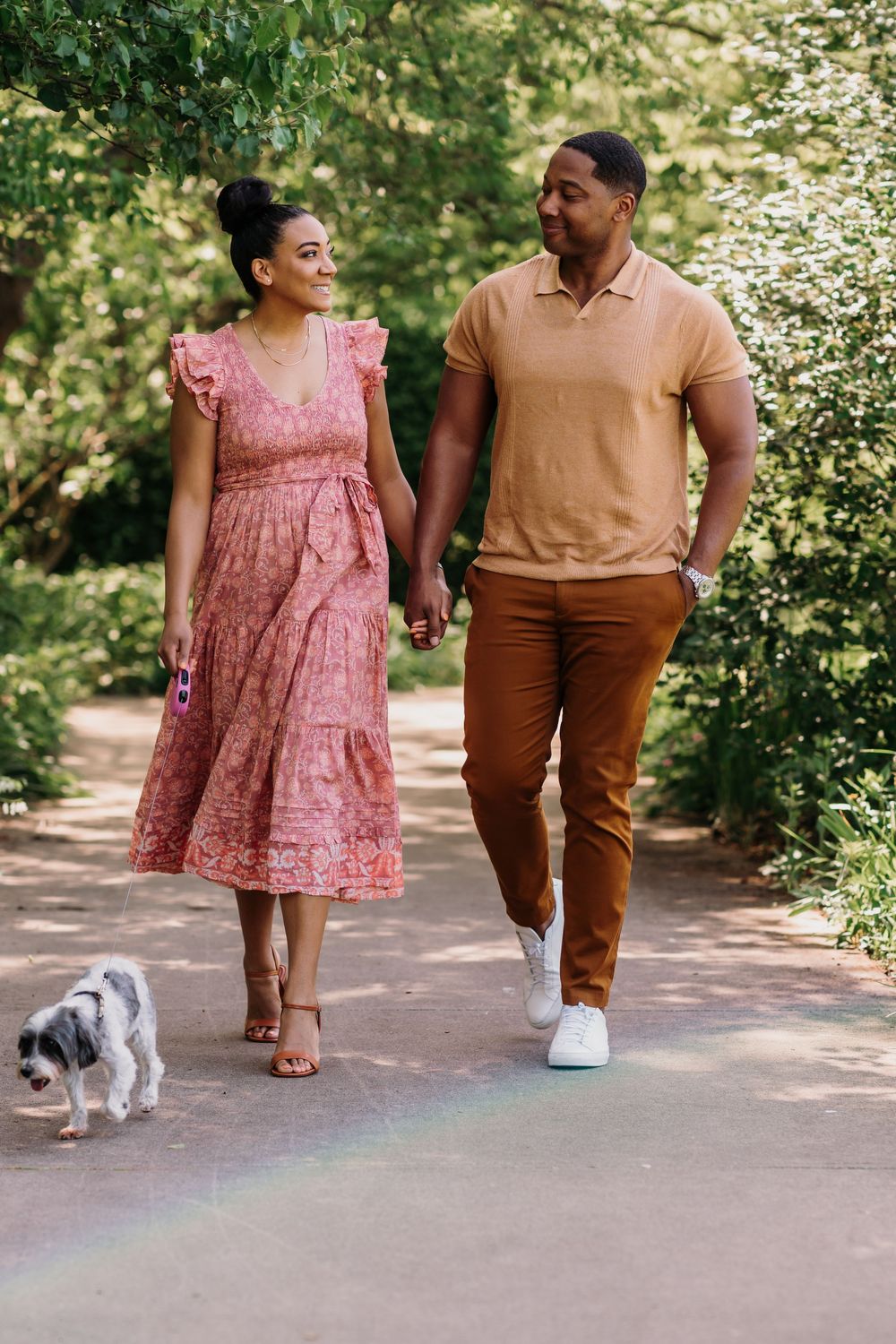 A couple in pink and brown outfits walk along a garden path with their small white dog. Couples Photographer Cincinnati