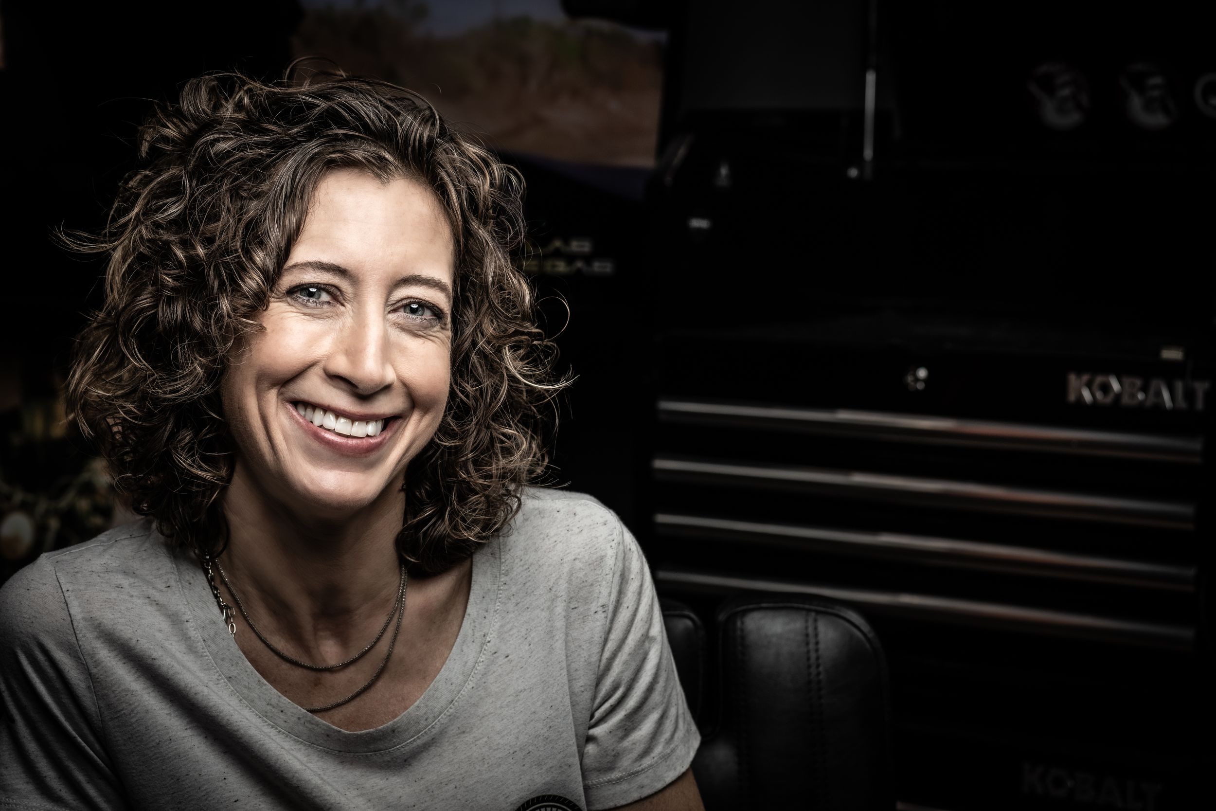 Person with curly hair wearing a grey shirt poses expressively against a dark background.