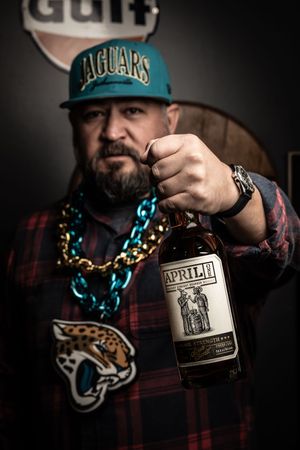 Person wearing Jaguars NFL gear holds up a bottle of Appleton Estate rum against dark background with turquoise accents.