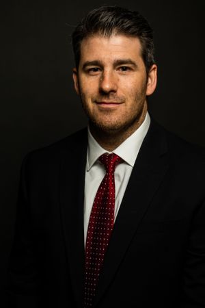 Professional portrait showing person in black suit with red patterned tie against dark background. Corporate Headshot