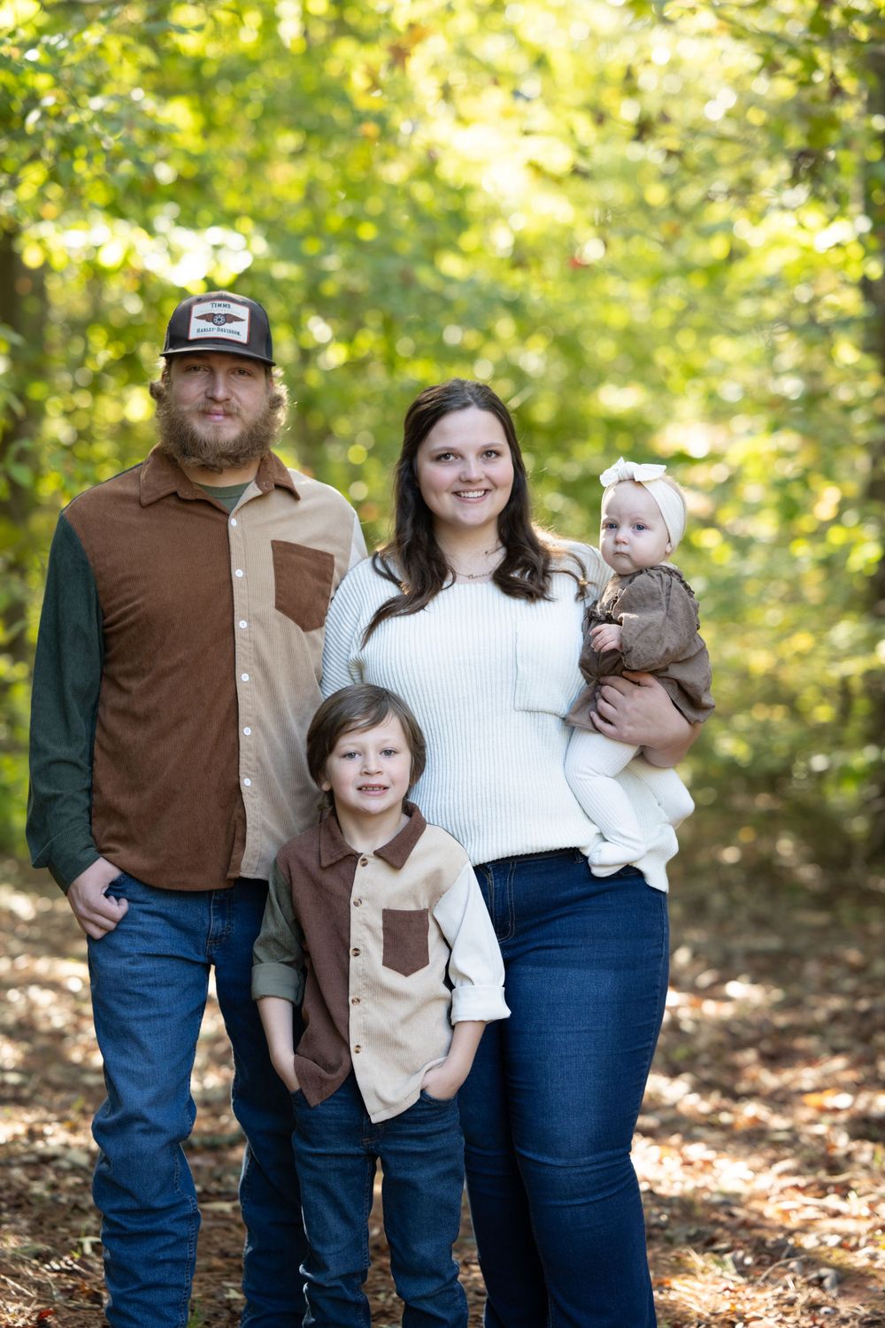 Family of four in coordinating brown and white outfits poses in sunlit autumn forest.