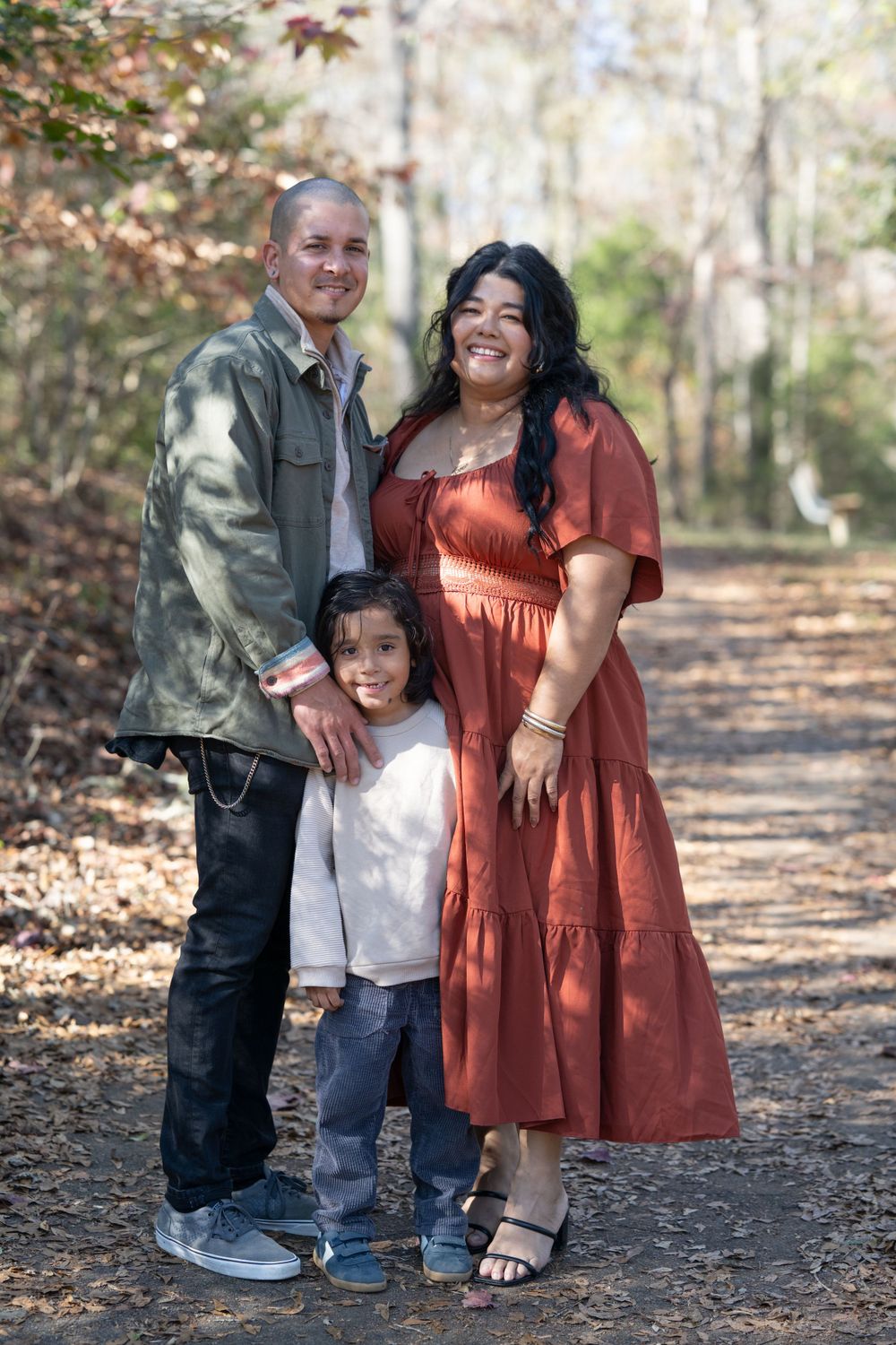 Family of three poses on wooded autumn trail in casual attire.