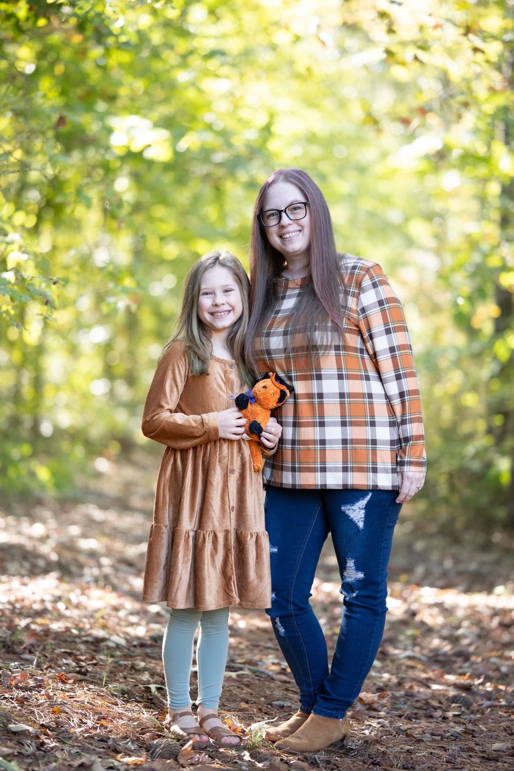 Mother and daughter in autumn clothing pose on leaf-covered trail holding small plush toy.