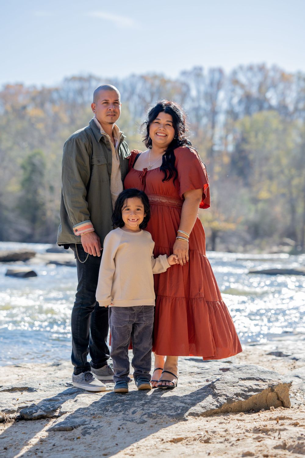 Family poses together on rocky riverbank with rushing water and autumn trees in background.
