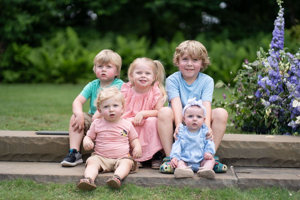 Group of young children sitting together on stone ledge with flowers in garden setting.