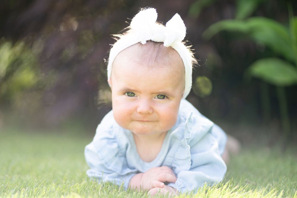 Baby in light blue outfit with white bow crawls on grass outdoors.
