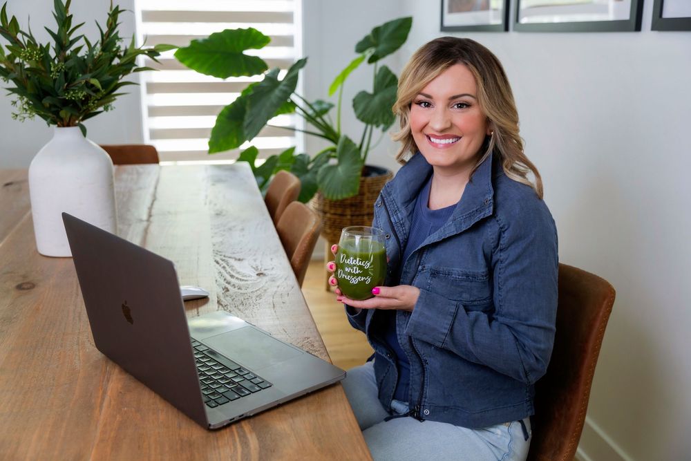 Person in navy blazer sitting at wooden desk with laptop and decorative plants in bright workspace.