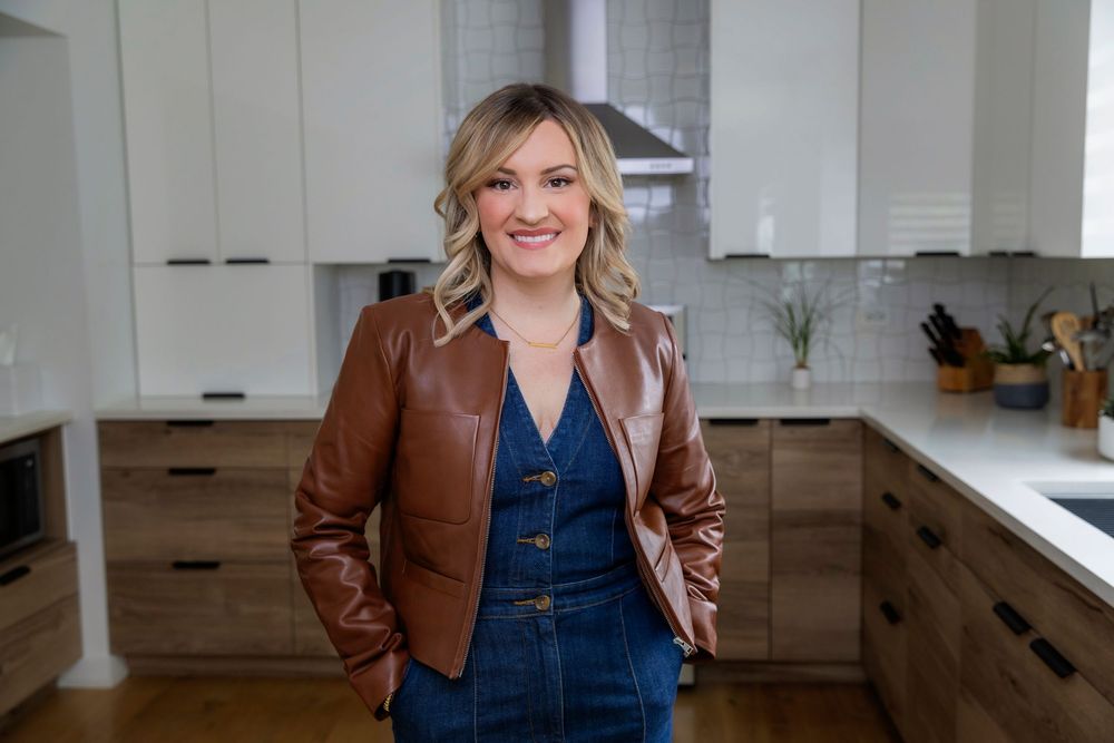 Woman in brown leather jacket and denim overalls stands smiling in modern kitchen with white countertops.