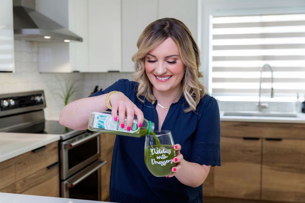 Someone in a navy blue top pours a green drink into a glass in a modern kitchen with wooden cabinets.
