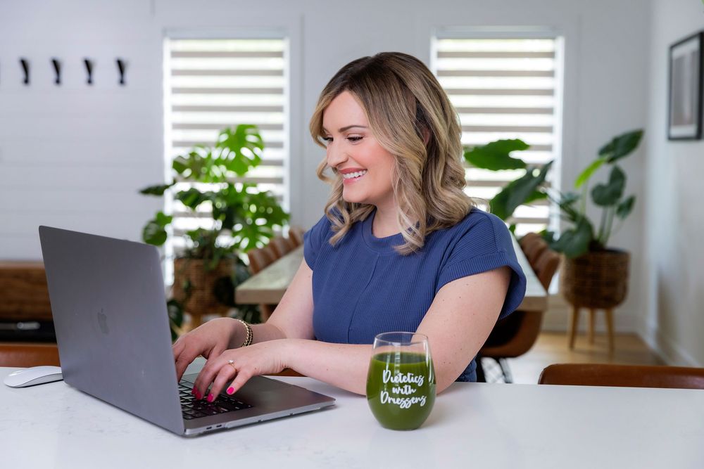 Remote worker at home office desk types on laptop with green plants and natural lighting in background.