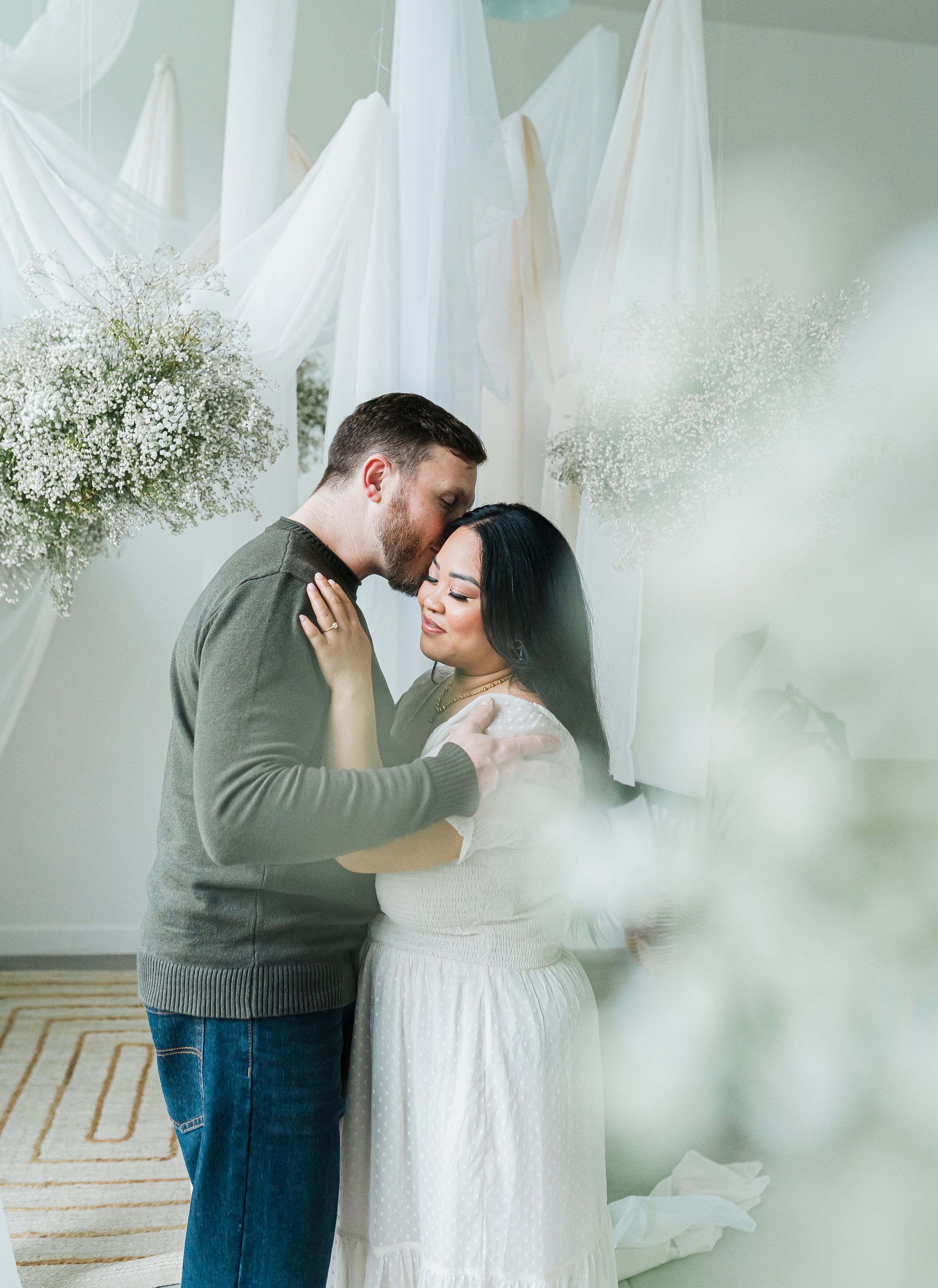 Romantic couple embracing in a dreamy white floral studio setting with flowing drapes and baby's breath arrangements.