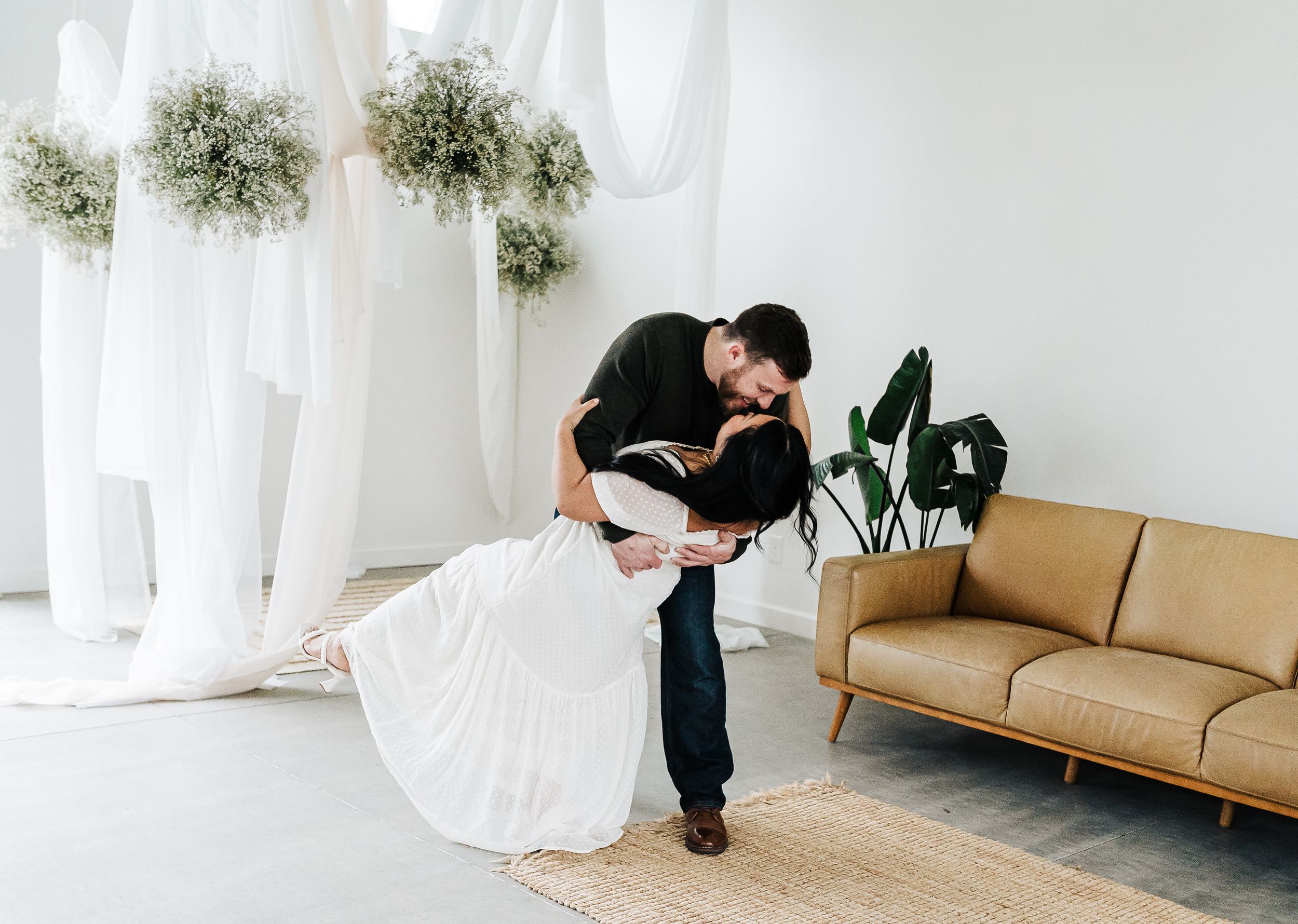Couple sharing a romantic dip kiss in a white decorated studio with floral arrangements and a tan leather sofa.