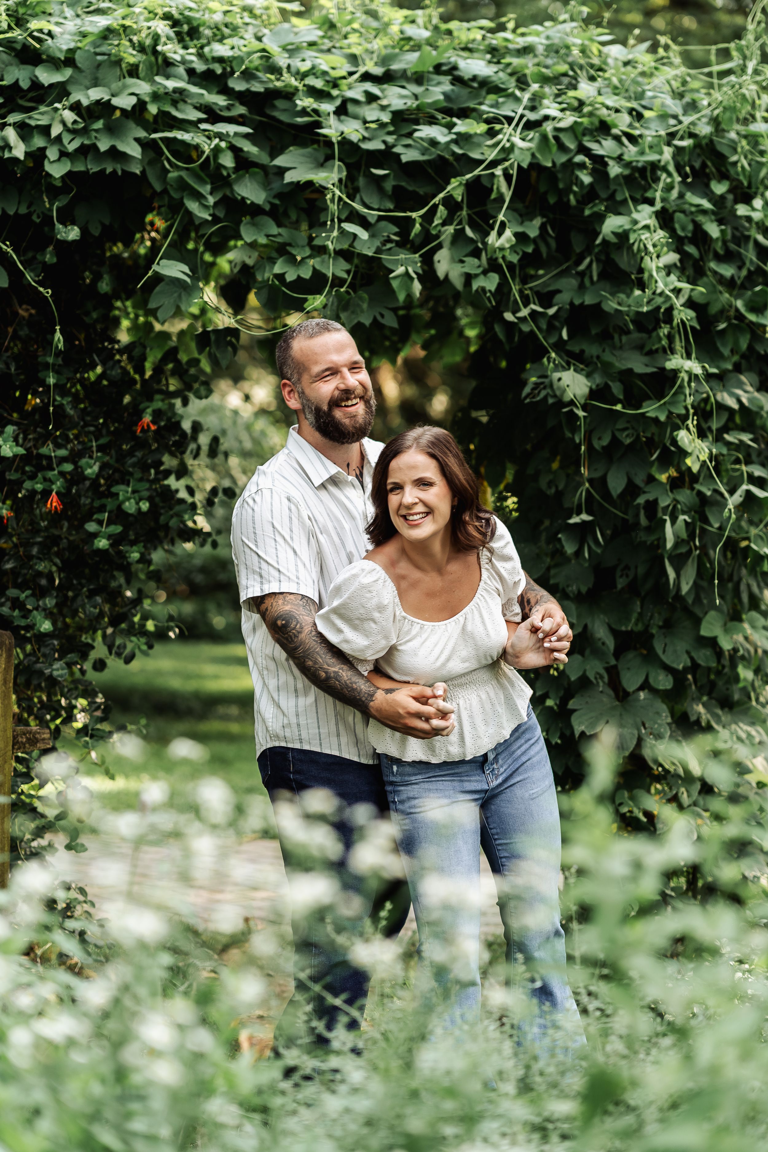 A smiling couple embraces in a lush green garden archway, the man with tattoos hugging the woman from behind.