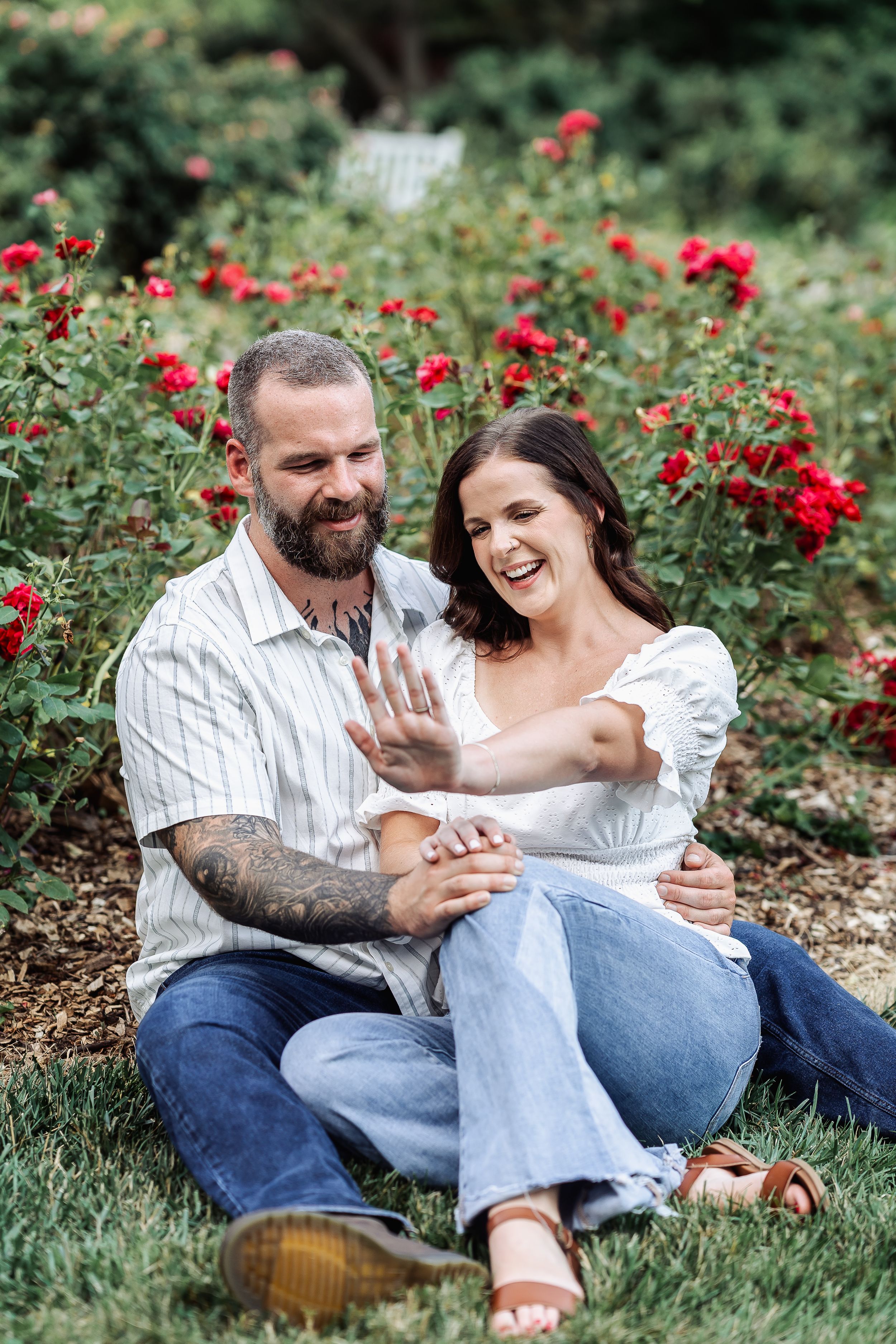 A happy couple sits in a rose garden, laughing as the woman shows off her engagement ring.