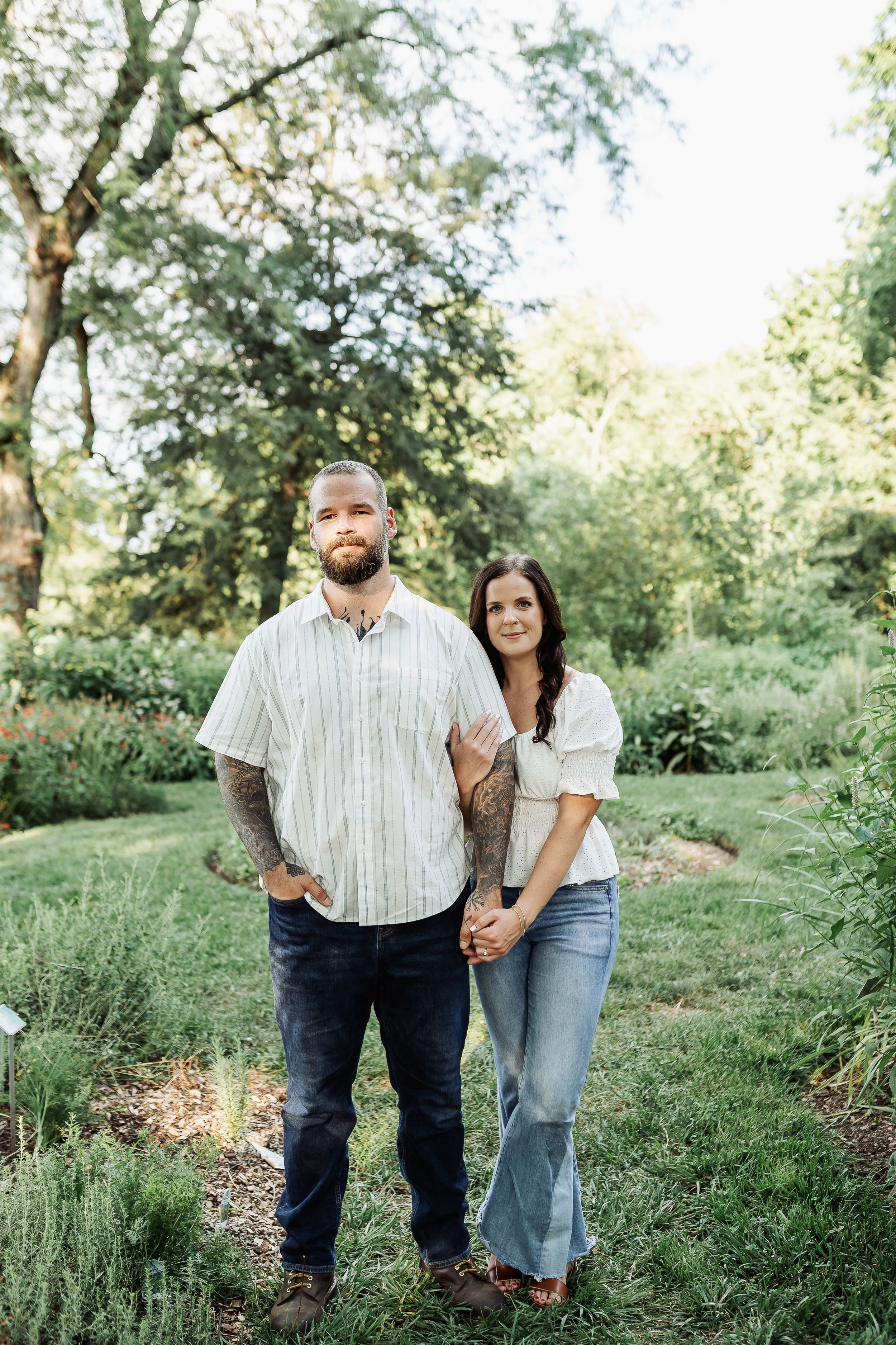 A couple holds hands in a lush green garden, man in striped shirt with tattoos, woman in white top and jeans.