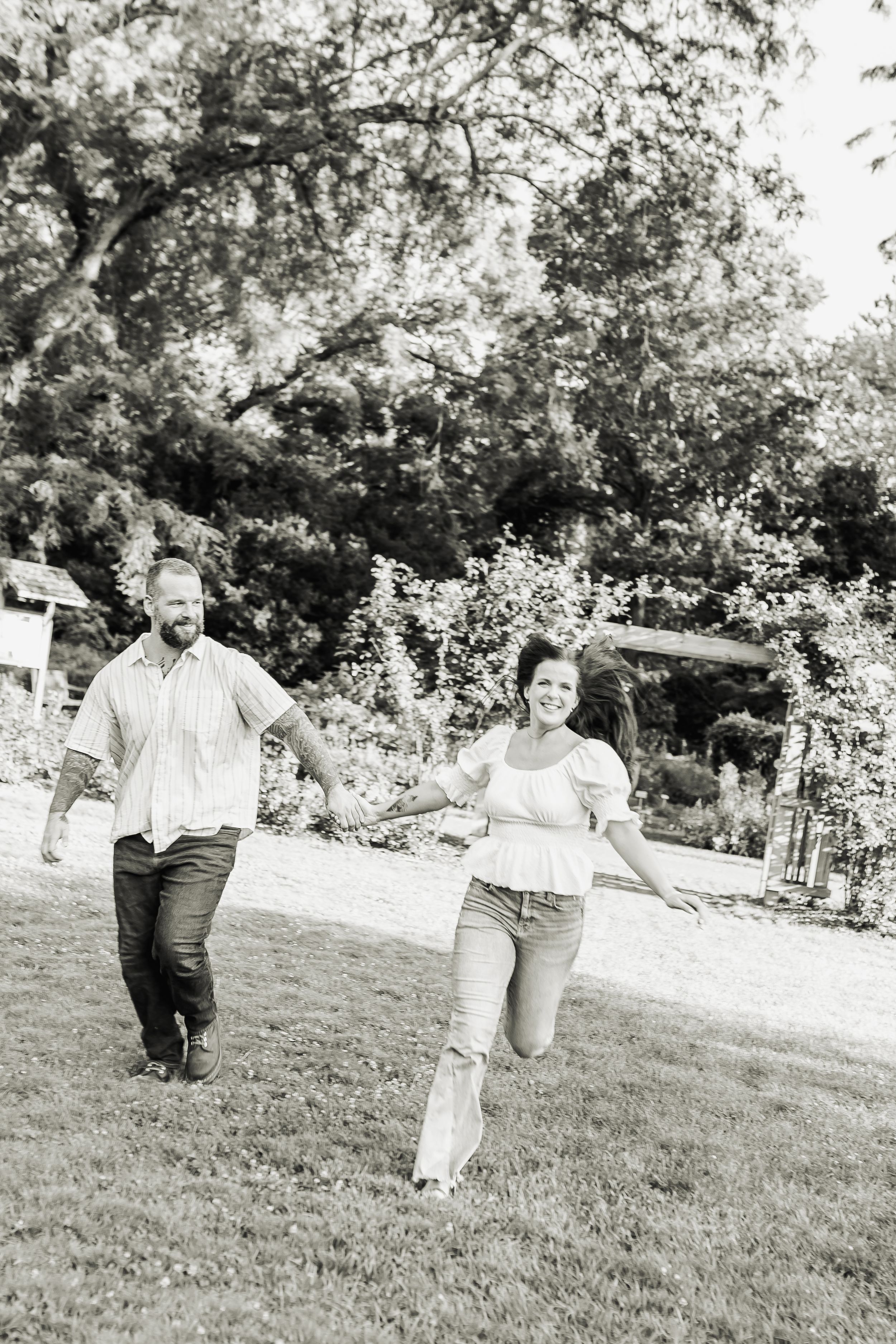 A joyful couple holds hands while running through a lush green garden in a black and white photo.