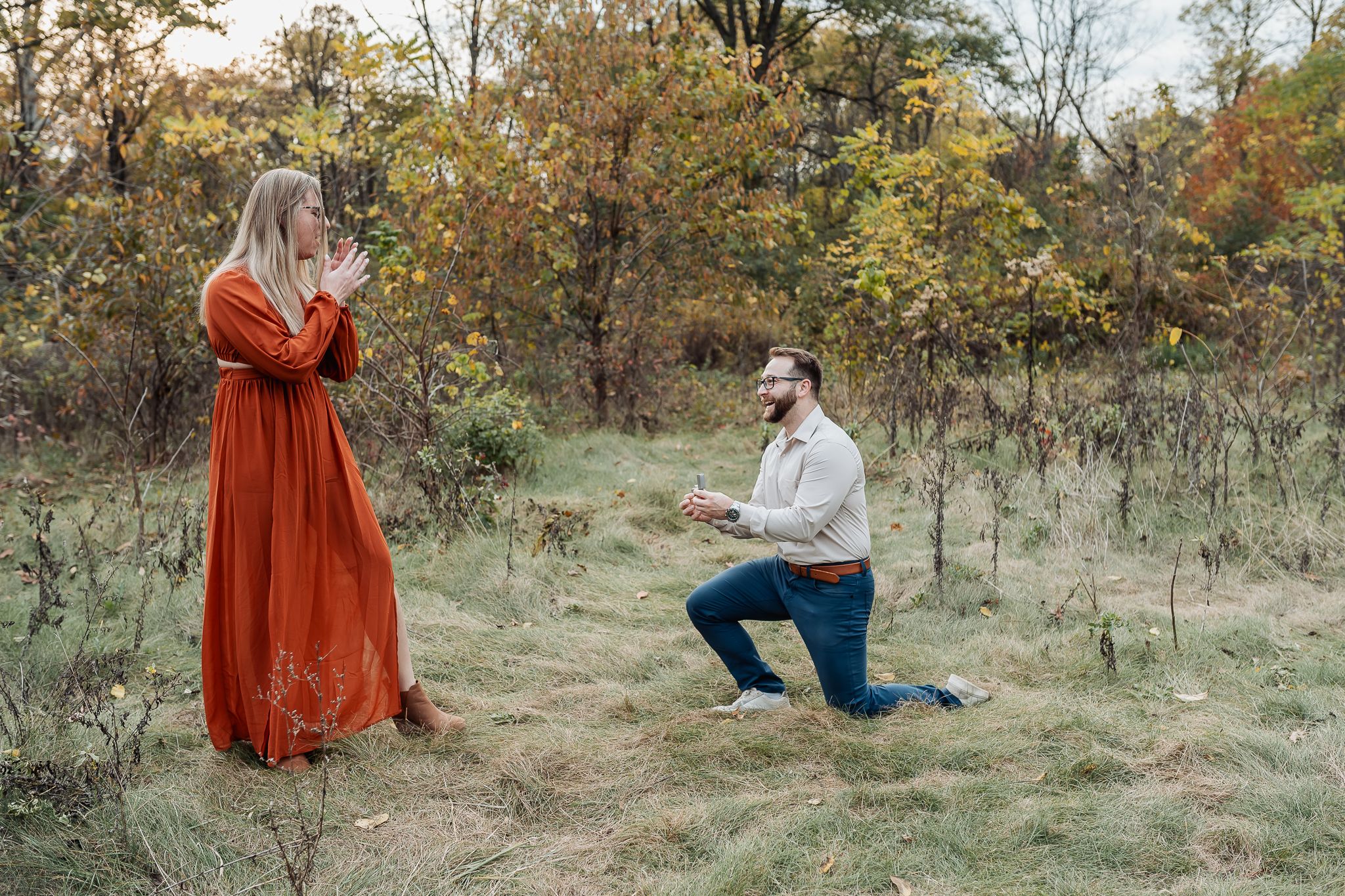 Man proposes on one knee to surprised woman in red dress outdoors in autumn forest setting.