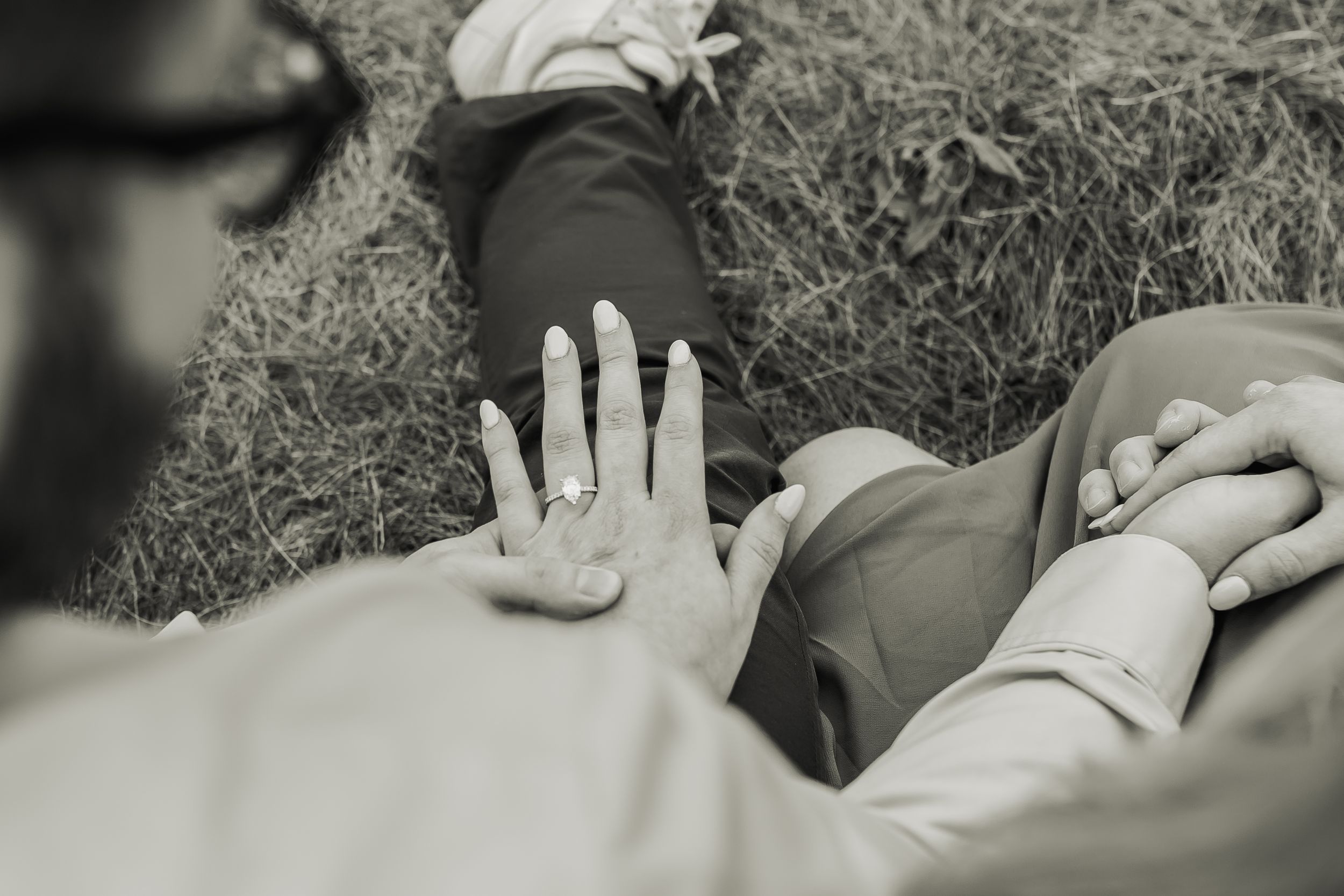 Black and white photo of a couple lying on grass, showcasing an engagement ring on the woman's hand.