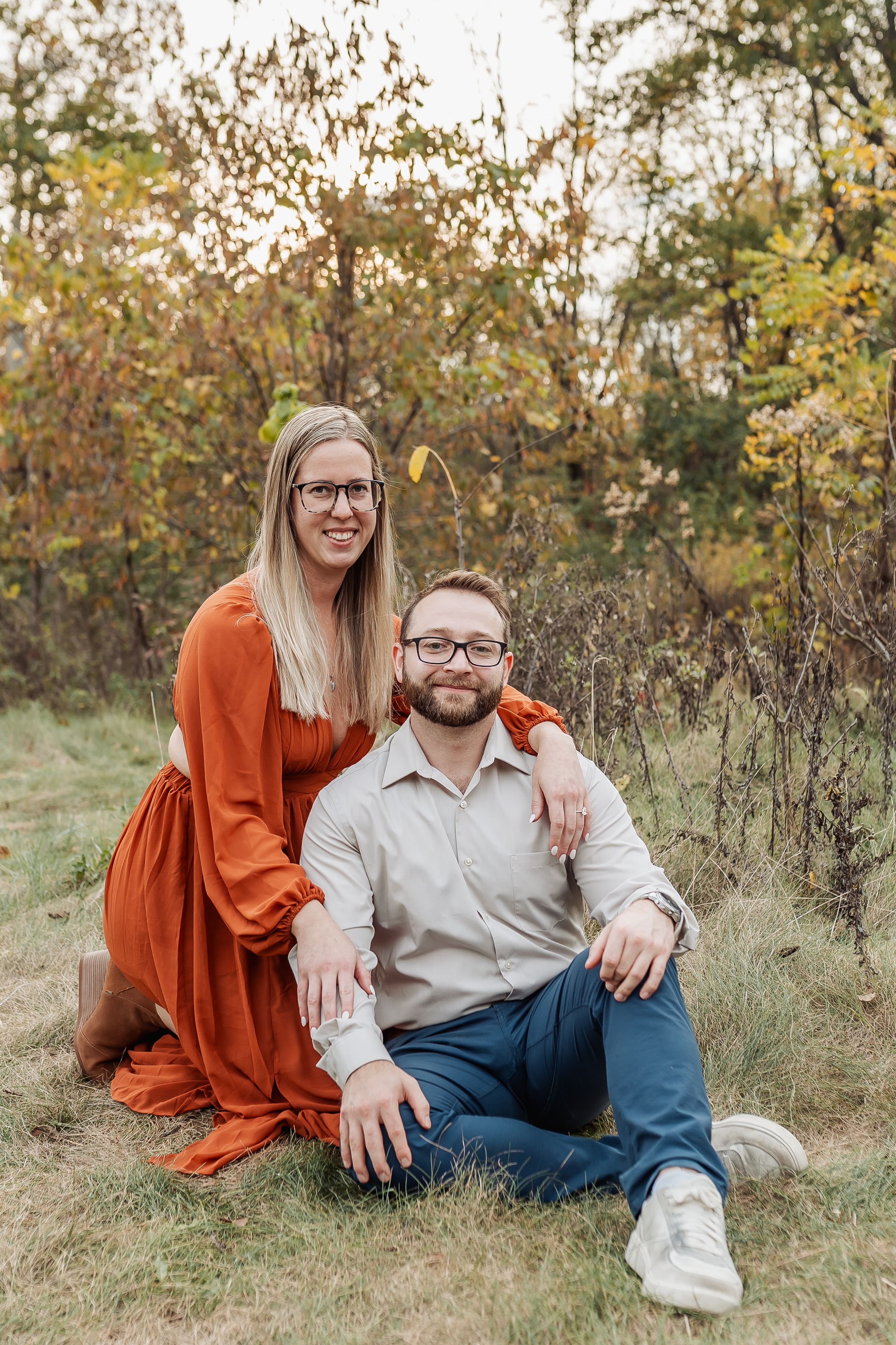 Couple posing outdoors in fall foliage, woman in rust dress standing behind seated man in gray shirt.