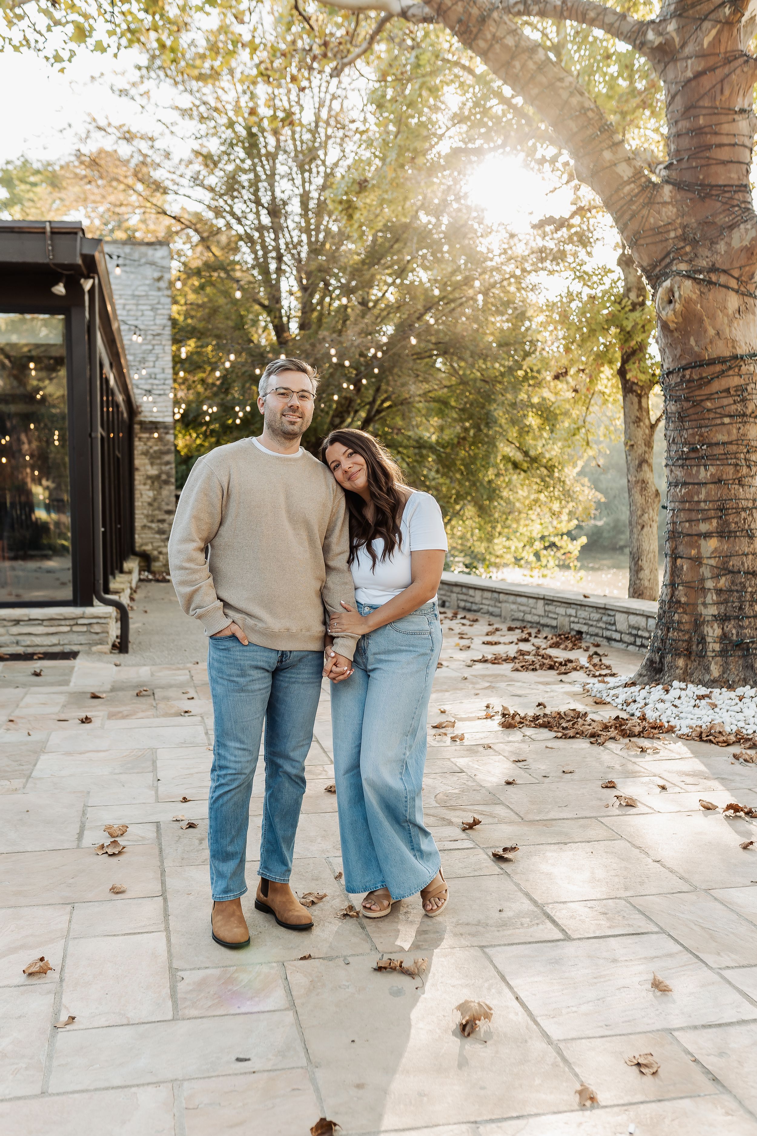A couple stands together on a stone patio surrounded by autumn trees, smiling warmly in casual jeans and sweaters.