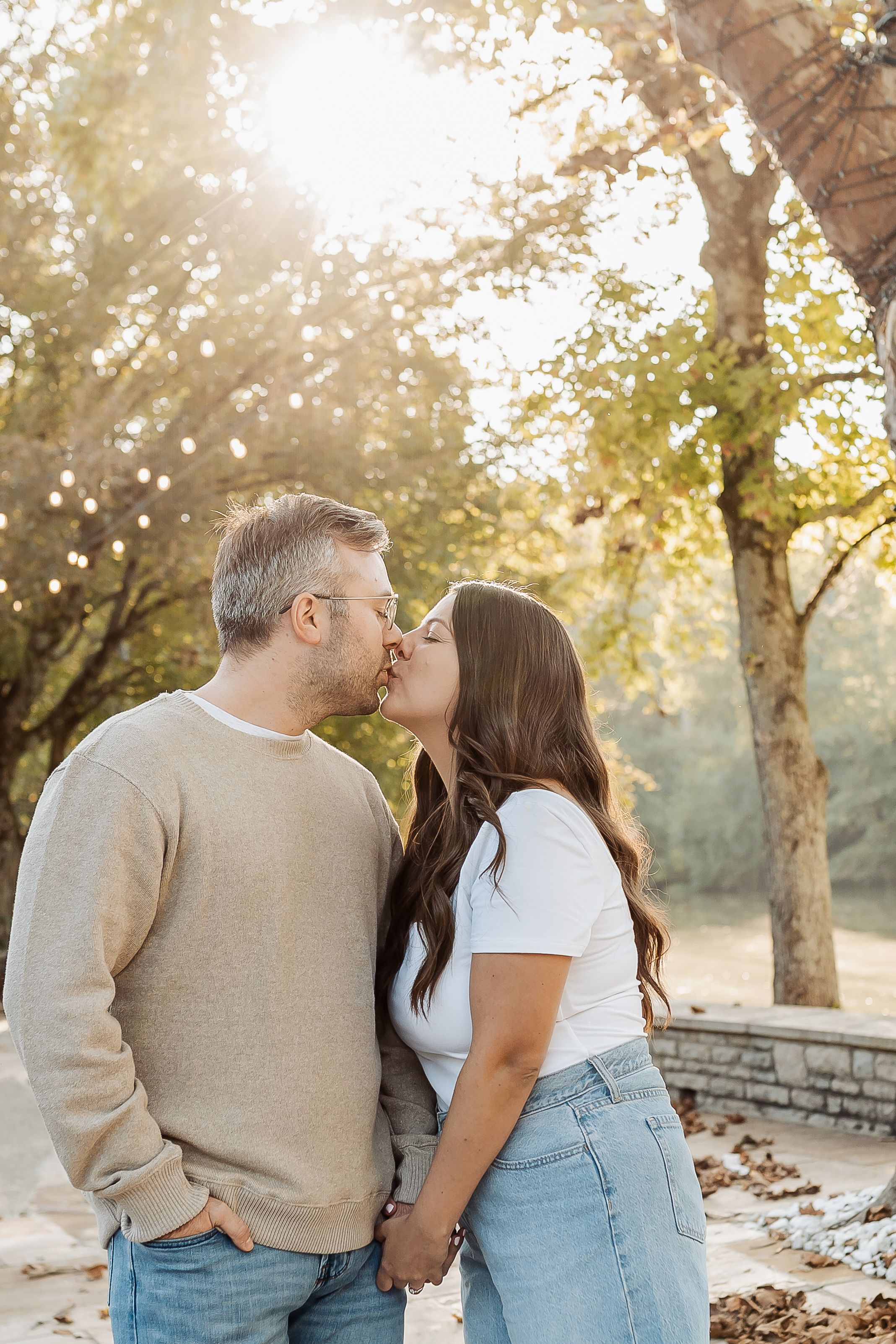 Couple sharing a romantic kiss while holding hands outdoors in golden autumn sunlight surrounded by trees.
